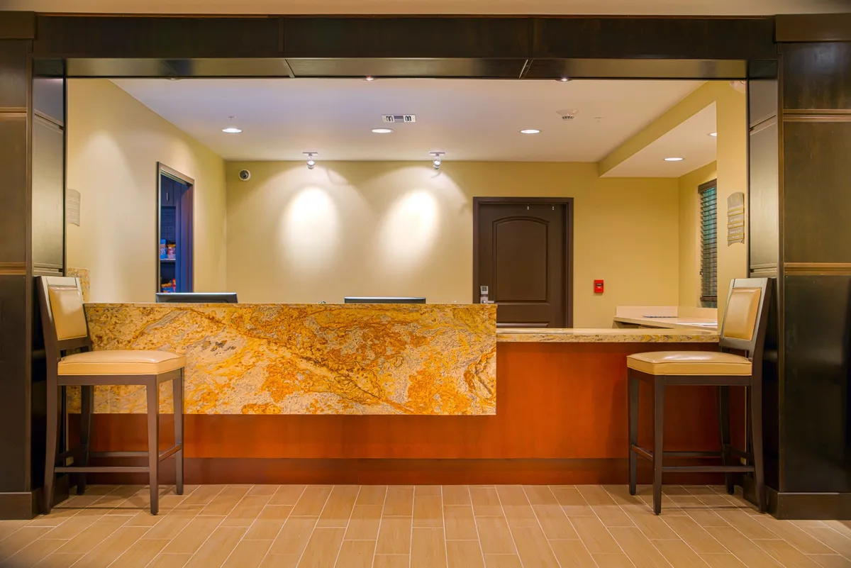 A modern hotel reception area featuring a granite front desk, stylish stools, and dark wooden accents. The scene includes guests checking in and staff offering assistance, creating an inviting first impression.