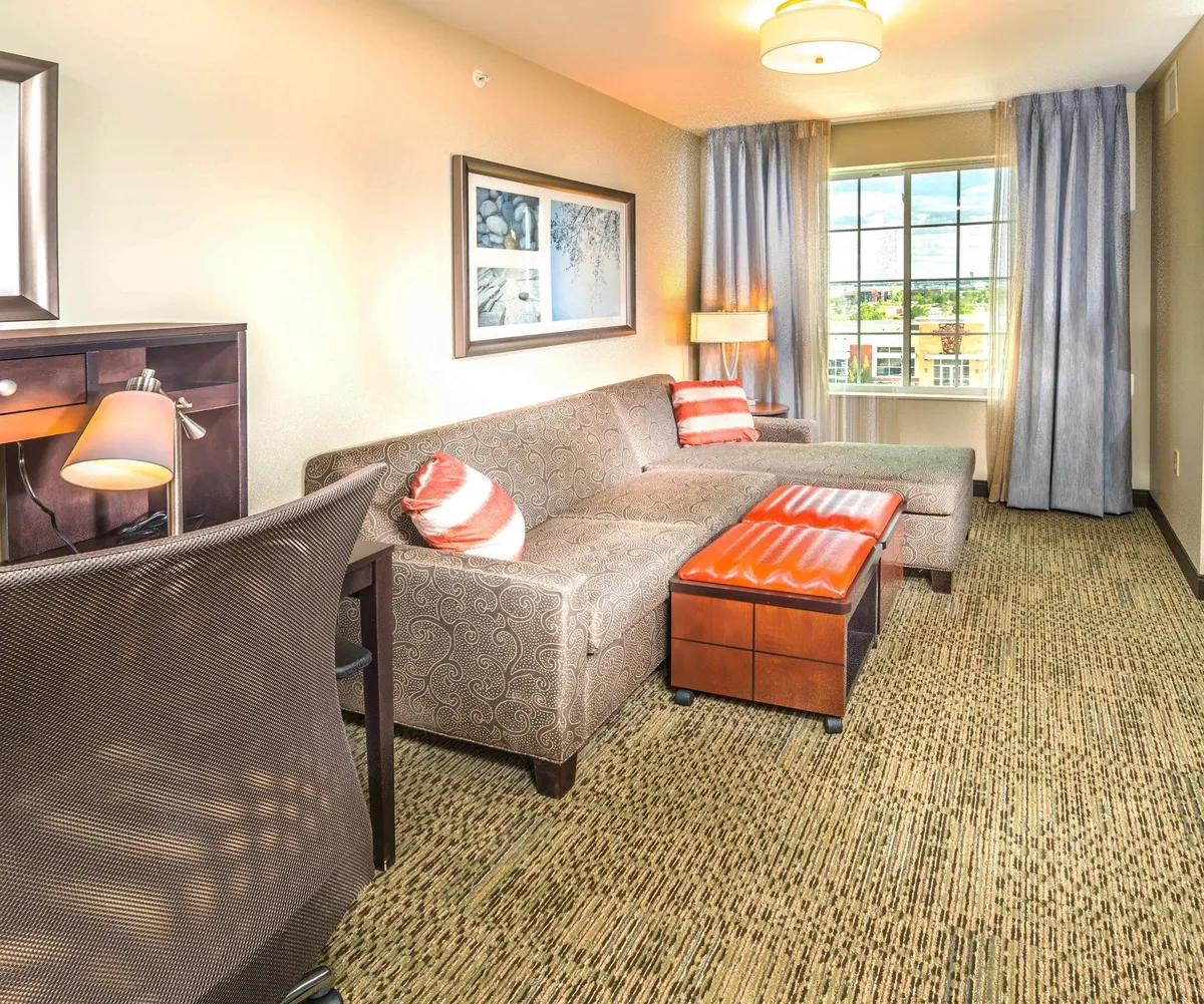 The image showcases a welcoming hotel suite interior with a plush sofa, a modern coffee table, a dedicated desk area, and a large window filling the room with natural light. The inviting space is perfect for morning coffee, productive work sessions, or relaxing after exploring Denver.