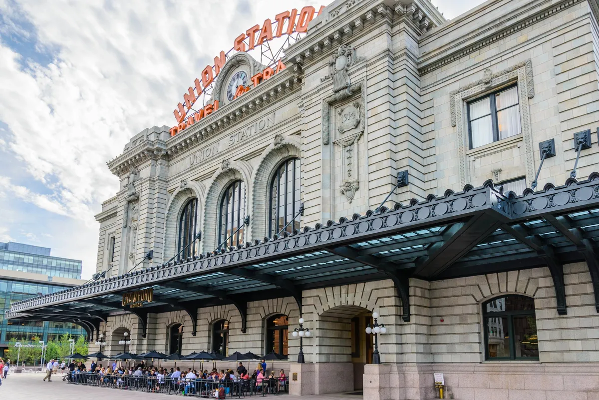The image showcases the charming exterior of Denver's historic Union Station, with its iconic sign, grand arched windows, and inviting outdoor seating area where people are dining and socializing. The lively atmosphere hints at local culture, historic architecture, and easy access to transportation.