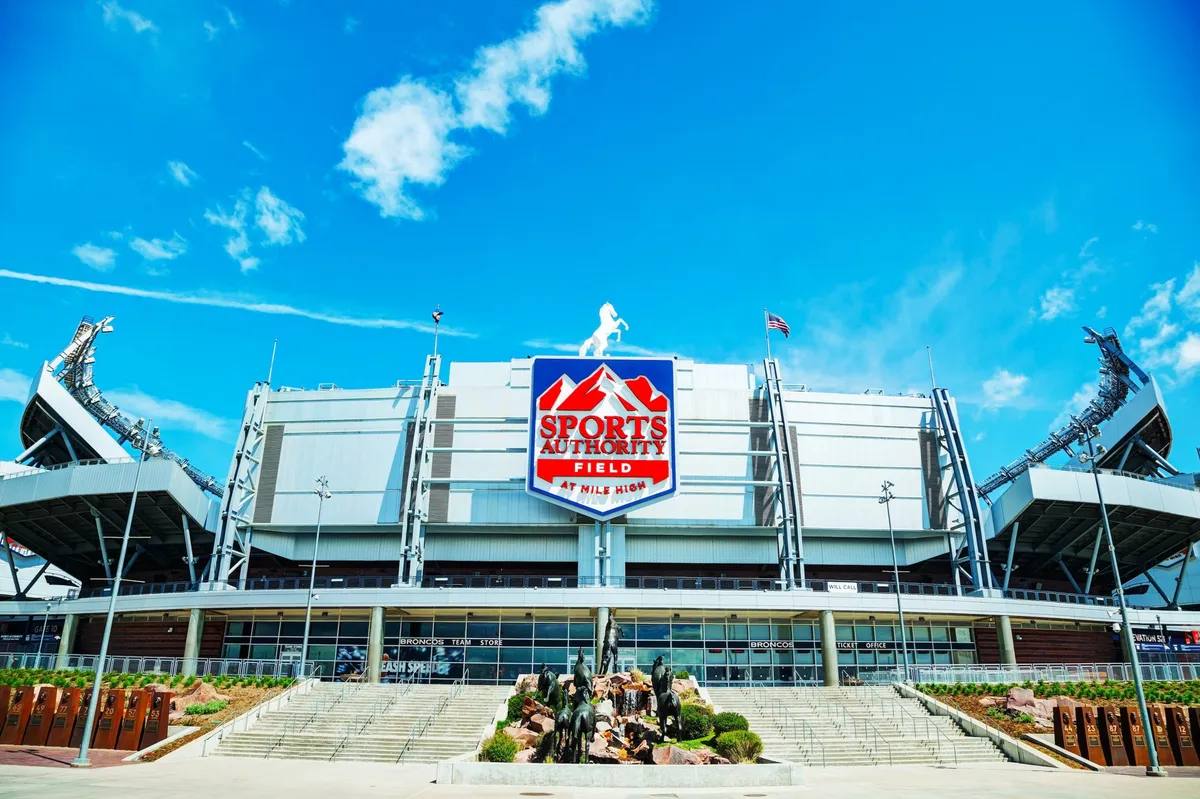 The image features the exterior of Sports Authority Field, showcasing the stadium's iconic structure and a prominent statue of football players. The scene is lively and dynamic, evoking the excitement of attending major sports events or community gatherings in Denver.