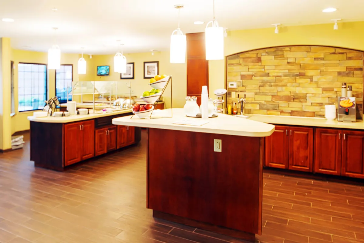A cozy dining area centered around a large island counter with a vibrant fruit display, complemented by warm lighting and a distinctive stone wall. The space feels inviting and designed for relaxed breakfasts, casual snacks, or enjoying coffee with friends. The central island provides a focal point for both dining and conversation, adding to the sense of warmth and community.