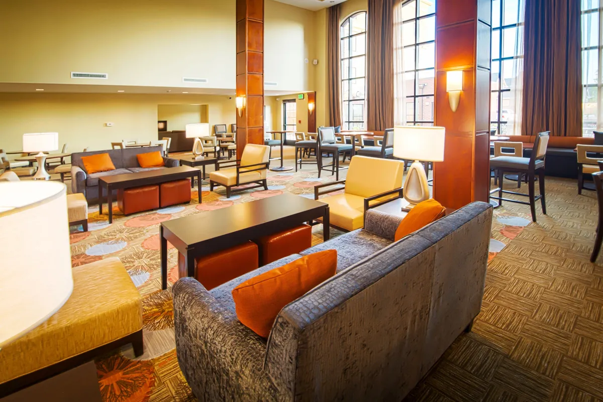 A spacious living area with comfortable seating, large windows letting in natural light, elegant lighting, and textured rugs. Guests can be seen reading, working on laptops, or enjoying conversations, highlighting the room's flexibility for both leisure and productivity.
