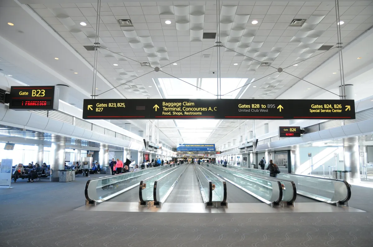 A bright, bustling interior space with a moving walkway, departure and arrival signs, and passengers either seated or traveling. The scene suggests a sense of movement and travel readiness, echoing the convenience of hotel living for travelers.