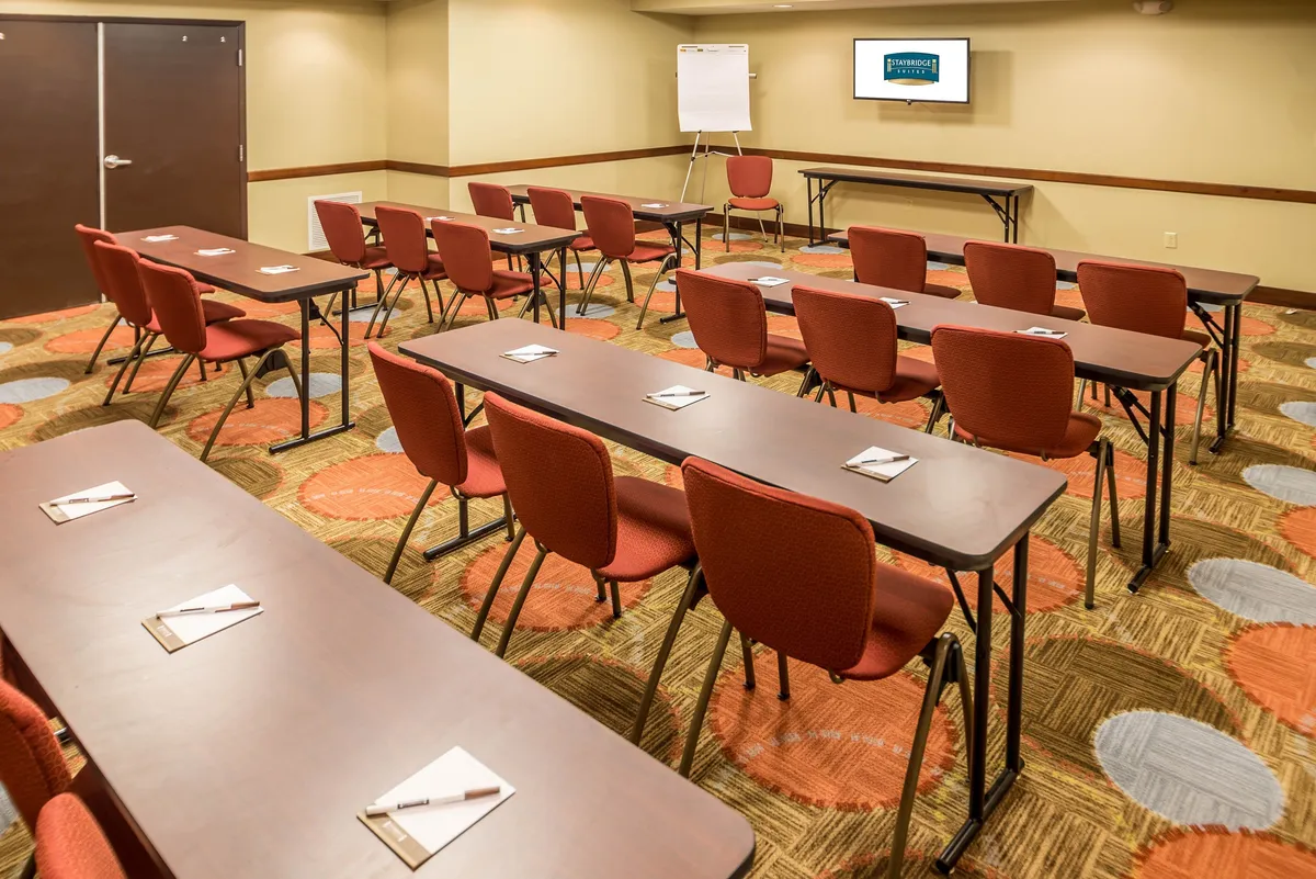 The image shows an interior meeting space with tables neatly arranged, red chairs, and a presentation screen. The room is well-lit and organized, suitable for gatherings, group activities, or collaborative sessions.