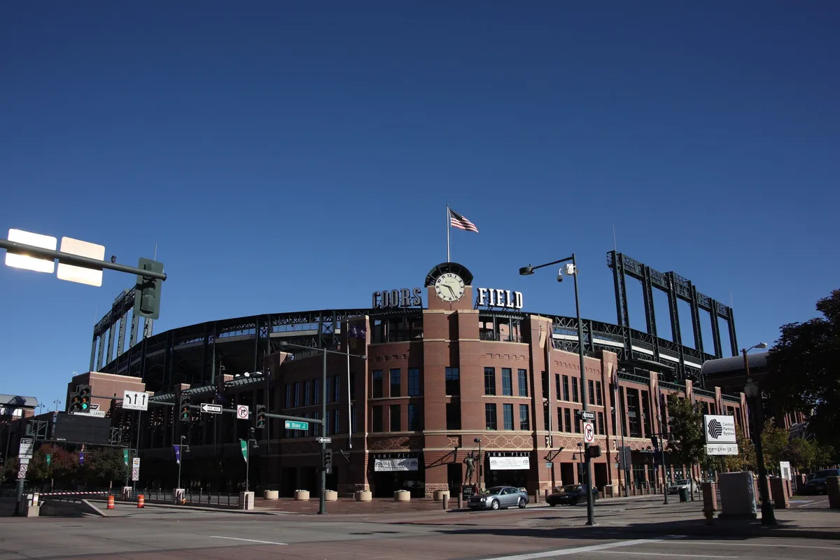This image features the recognizable Coors Field sign, an American flag waving proudly, and the classic brick facade of the stadium. The bustling exterior captures the energy of a game day, with spectators likely heading in for a Rockies baseball game or participating in pre-game festivities.