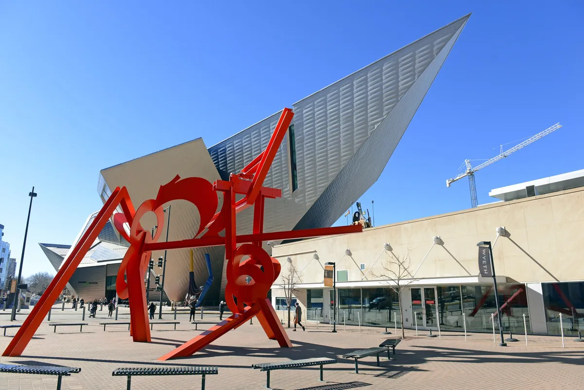 A striking modern sculpture stands before a contemporary architectural structure, surrounded by benches where visitors are relaxing and taking photos. The scene evokes Denver’s artistic and cultural side, inviting exploration of public art and community spaces.