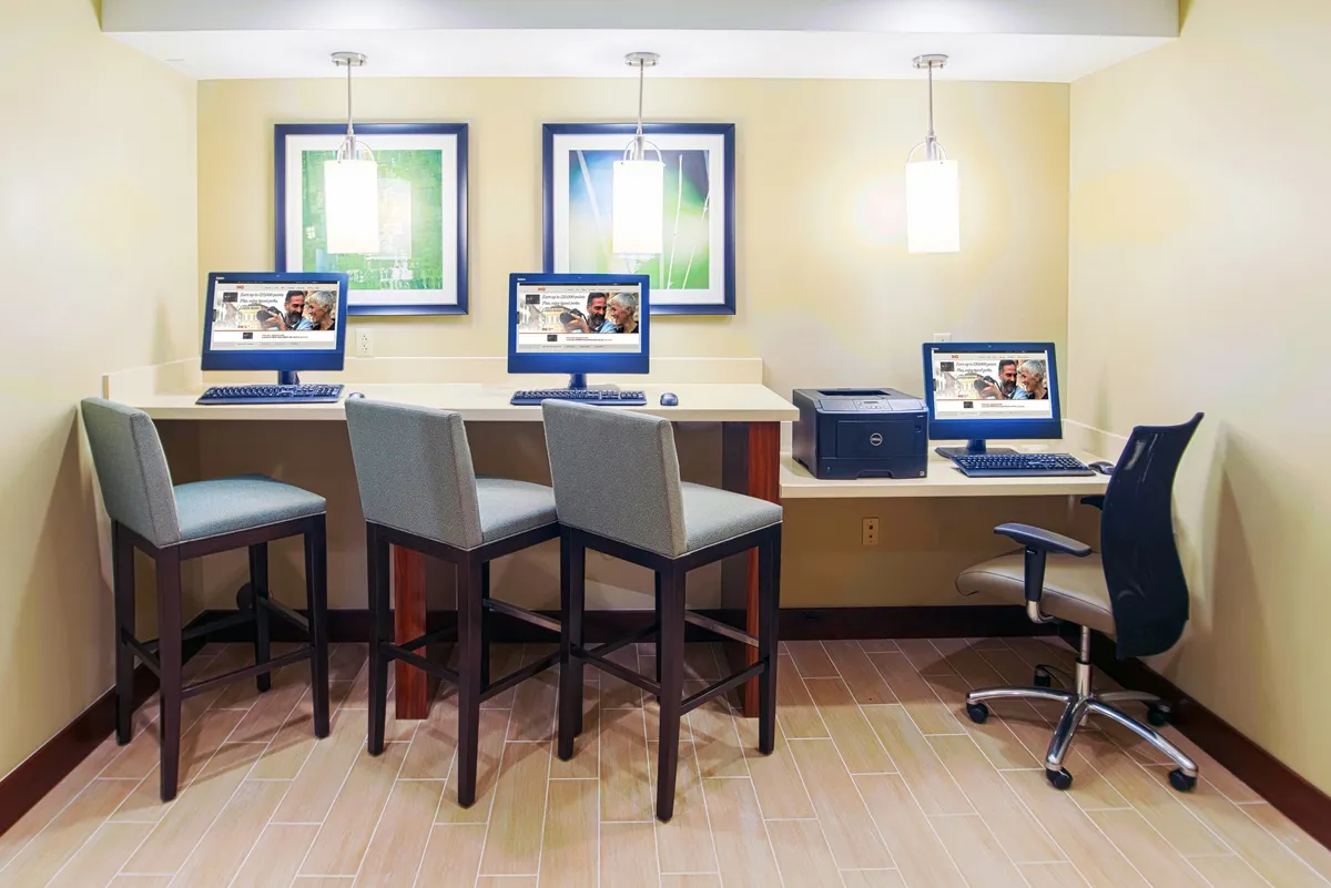 A well-lit business center featuring a row of computers on a countertop with modern bar stools, complemented by decorative artwork. The setup is designed for productivity, with ample workspace and easy access to technology for working, printing, or casual meetings.